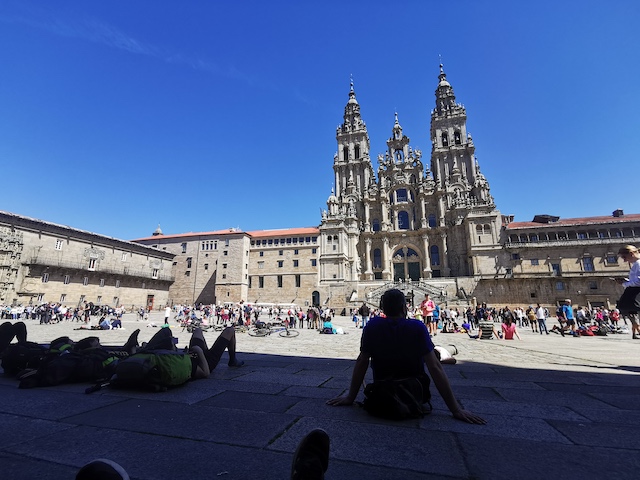 威厳のある大聖堂と、その前の広場の地面に座り込んだり横たわったりしている大勢の巡礼者