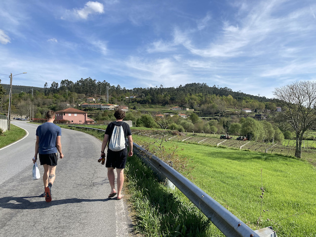 緑が綺麗なポルトガルの村の道路を、ビール瓶とビニール袋を持って歩く若者2人