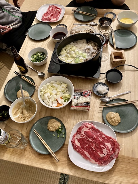 テーブルに所狭しと並べられた食事（野菜の入った鍋、霜降り肉、タレ、うどんなど）