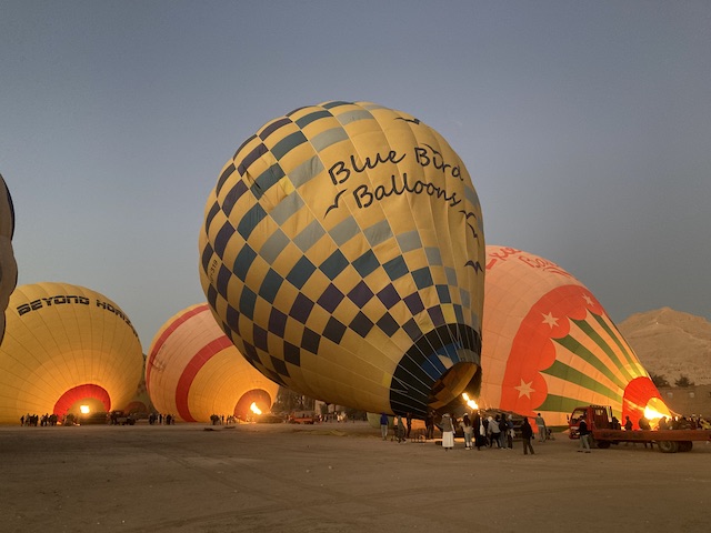 薄暗い空の下、地面に置かれて斜めに立ち上がろうとしている複数の気球と、その下でバーナーを操作している人たち