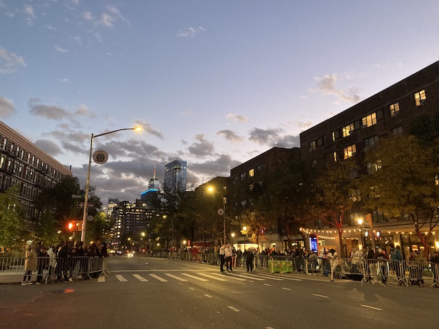 薄闇の中で街灯に照らされながら、フェンスの外でパレードの始まりを待っている人々